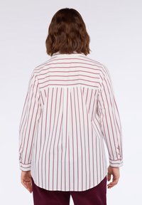Camicia bianca a maniche lunghe con sottili strisce verticali color bordeaux. Presenta un yoke posteriore e un orlo curvo. Polsini con bottoni e piccole aperture a fessura.