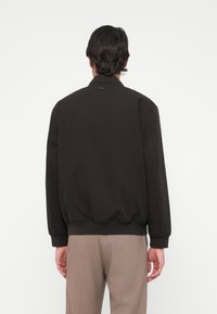 Must muströöt jakk, mis on valmistatud siledast kangast, ribitud mansettide ja alumise äärega. Lacoste logo onemplodeeritud taga kaelas.