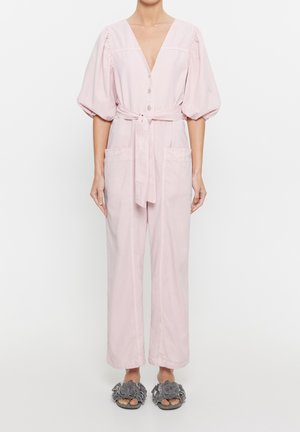 Kvinde iført lyserød knaplukket jumpsuit med puffede ærmer og bindebånd i taljen, kombineret med grå slip-on sandaler med blomsterprint.