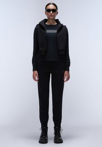 Zwarte sweatshirt met een capuchon en logo-detail, gecombineerd met zwarte tapering joggers en zwarte veterschoenen. Glad materiaal, ontspannen pasvorm.