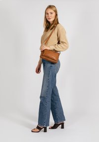 Brun läder crossover-väska med slät textur, rektangulär form och justerbar rem. Matchas med en beige tröja och blå jeans.
