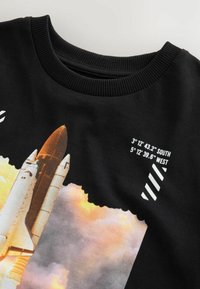 Zwarte T-shirt met een grafische afbeelding van een spaceshuttle die wordt gelanceerd tegen een achtergrond van wolken. Bevat coördinaten in witte tekst en een geribde halslijn.