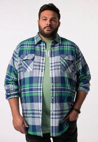 Camicia flanella a quadri nei toni di blu, verde e grigio. Presenta due tasche sul petto con chiusure a scatto e una chiusura frontale con bottoni. Il tessuto ha una consistenza morbida.