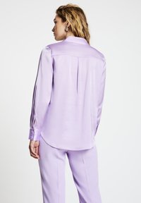 Woman with wavy blond hair wears shiny lavender blouse with long sleeves and matching lavender pants, facing away against white background.