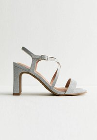Sandale à talon haut argentée avec un design de sangle texturée, bout ouvert, boucle réglable et un talon bloc. Semelle intérieure lisse et semelle extérieure de couleur claire.