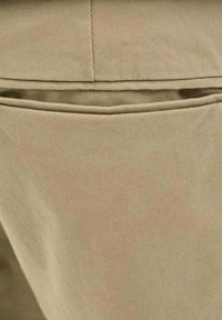 Tessuto beige con una consistenza liscia, dotato di un dettaglio a tasca orizzontale in vita. Il tessuto è resistente e strutturato.