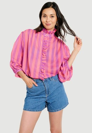 Jeune femme tirant ses cheveux, portant un chemisier volanté à rayures roses et violettes et un short en jean bleu, debout devant un fond clair uni.