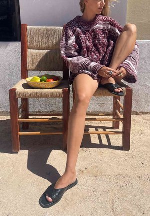 Frau in gemustertem Pullover sitzt draußen auf einem Holzstuhl, ein Bein übereinandergeschlagen, neben einem anderen Stuhl mit einer Schale Obst.