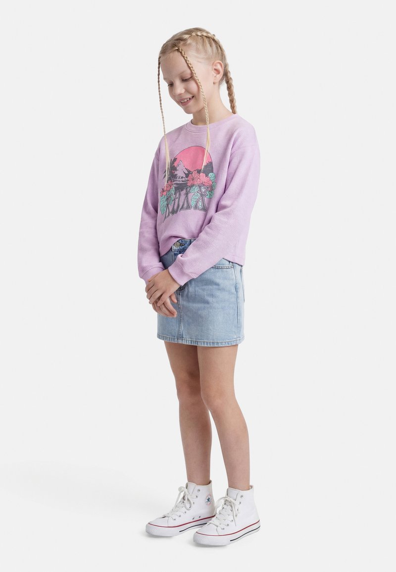 Mladá dívka s blond copánky, oblečená do fialové mikiny s tropickým motivem, džínové sukně a bílých vysokých tenisek, stojí a usmívá se.