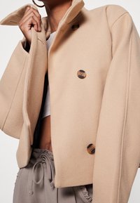 Personne portant un manteau en laine beige avec de gros boutons, tenant le col, associée à un pantalon taupe à cordon et un haut blanc court.