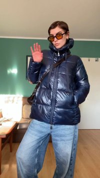Casaco puffer azul marinho com textura brilhante, fecho com zip, cordões ajustáveis e bolsos laterais, combinado com jeans azul claro e óculos de sol.