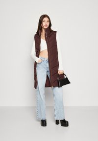 Gilet sans manches matelassé bordeaux avec un grand col et une fermeture éclair dorée. Associe à un top blanc court, un jean délavé, un sac à main noir et des chaussures à plateforme.