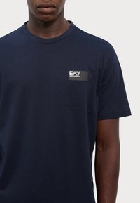 Navy zila kokvilnas t-kreklu ar apaļu apkakli, īsām piedurknēm un mazu taisnstūrveida EA7 logotipa uzlika kreisajā krūtī.