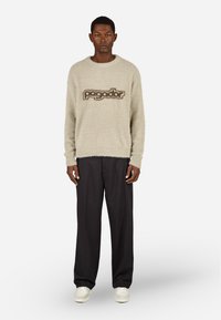 Bežs džemperis ar apaļu kaklu, izgatavots no pūkaina auduma, ar tumši brūnu "pagador" logo. Kombinēts ar brīviem melniem biksēm un baltām sporta apaviem.