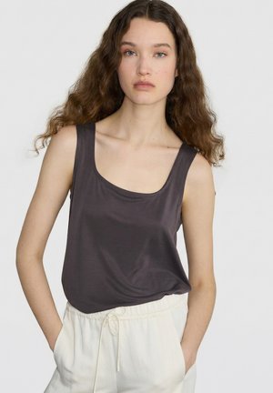 Jonge vrouw met lang krullend haar die een donkergrijs mouwloos topje en witte broek met trekkoord draagt, staand tegen een effen achtergrond.