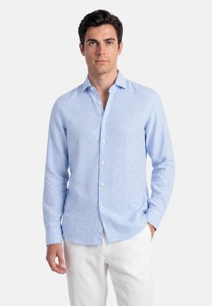 Uomo con capelli scuri che indossa una camicia azzurro chiaro a maniche lunghe con bottoni e pantaloni bianchi, in piedi con una mano in tasca.