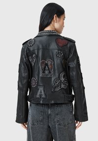 Woman wearing black studded leather jacket decorated with various embroidered patches, paired with faded black denim jeans, facing away.