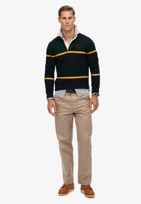 Blauw rugbyshirt met lange mouwen, voorzien van groene en gele strepen en een witte kraag. Beigekleurige chinos en bruine schoenen maken de outfit compleet.