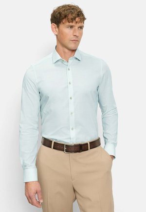 Mann mit lockigem Haar, der ein hellblaues Hemd mit Knöpfen und beige Hosen trägt, mit einer Hand in der Tasche stehend und nach rechts blickend.