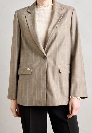Blazer beige a rayas con un solo botón, bolsillos con solapa y pequeñas iniciales doradas "M.M.", usado sobre una blusa de punto color crema con puños fruncidos.