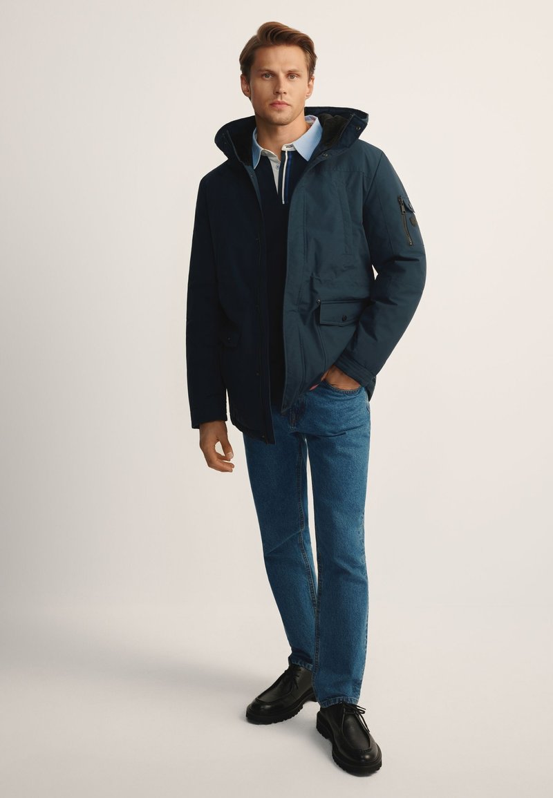 Mężczyzna w granatowej, zimowej kurtce z kapturem, jasnoniebieskiej koszuli z kołnierzykiem, niebieskich dżinsach i czarnych butach stoi na tle jednolitego tła.