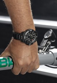 Melna analoga pulksteņi uz rokas, ar apaļu ciparnīcu, kontrastējošām baltām un sarkanm krāsām, un matētu apdari. Roka turpelē pelēku stūri ar zaļu uzrakstu.