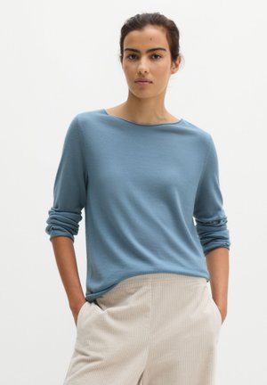 Frau trägt ein hellblaues Langarmshirt mit hochgekrempelten Ärmeln und beige Cordhosen mit den Händen in den Taschen vor einfarbigem Hintergrund.