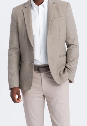 Hombre que lleva una chaqueta beige, camisa blanca, pantalones beige claro y cinturón gris con una mano en el bolsillo.