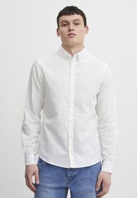 Camisa blanca de manga larga y con botones, confeccionada en una tela ligera, con un cuello clásico y botones amarillos, combinada con unos jeans de mezclilla azules.