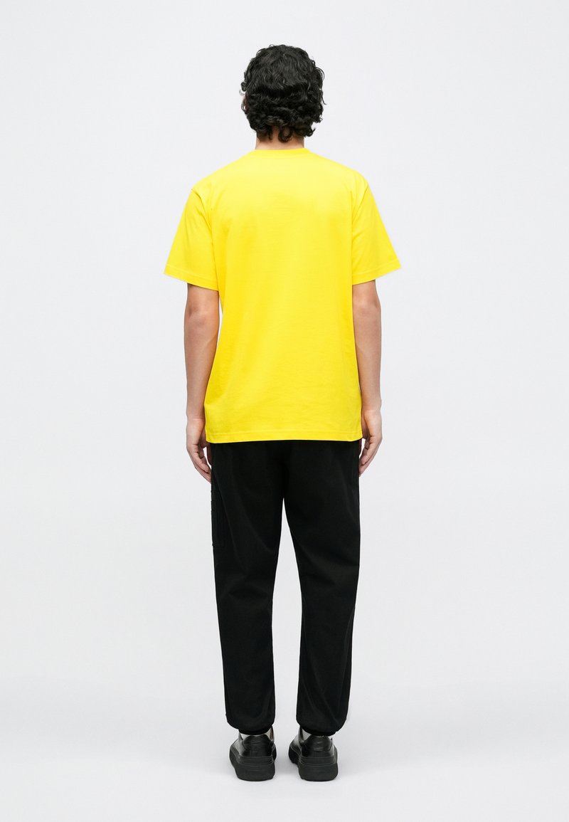 Gul bomuld t-shirt med korte ærmer, kombineret med sorte bukser. T-shirten har rund hals og en afslappet pasform.