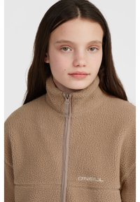Beige fleece jacket with high collar and zipper closure, featuring textured fabric and an embroidered logo in white on the chest.