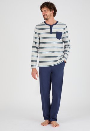Homme portant une chemise henley rayée à manches longues avec une poche poitrine et un pantalon bleu marine uni, debout pieds nus et regardant sur le côté.