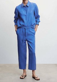 Blauw linnen overhemd met knoopsluiting en opgerolde mouwen, gecombineerd met blauwe broek met trekkoord. Standaard pasvorm met een ontspannen silhouet en platte sandalen.