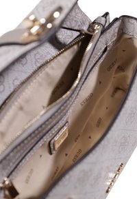Borsa a tracolla grigia con motivo e hardware dorato, dotata di chiusura con cerniera e interno con fodera beige e logo "GUESS" stampato ripetutamente.