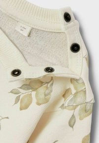 Off-white džemperis ar ziedu rakstu, kas rotāts ar dzeltenām citronām; tam ir melni spiedpogu pogas pie apkakles un teksturēts iekšpuse.