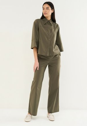 Femme debout portant une chemise à boutons vert olive à larges manches et un pantalon assorti, chaussée de beige, sur fond blanc uni.