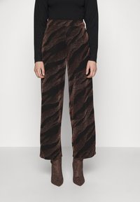 Pantalons amples marron avec un motif texturé en zigzag dans une teinte marron plus foncée, portés avec un haut ajusté noir à manches longues et des bottines à paillettes marron.