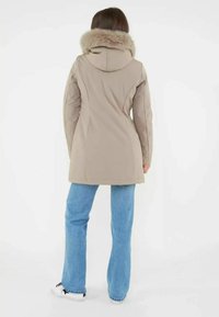 Beżowy płaszcz z futrzanym kołnierzem, dopasowany krój, długie rękawy i detal szwu z tyłu. W połączeniu z jasnoniebieskimi dżinsami i trampkami.