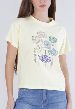 Femme portant un t-shirt jaune pâle avec des imprimés floraux multicolores et des mots manuscrits, associé à un jean bleu sur un fond uni.