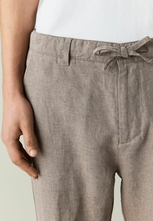 Prim-plan cu o persoană care poartă pantaloni bej din in, cu talie legată și șlițe pentru curea, asortați cu o cămașă albă.