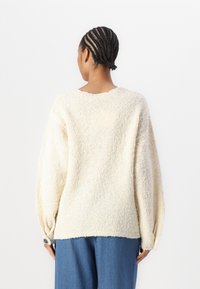Cream-colored, textured sweater with a loose fit, dropped shoulders, and buttoned cuffs, paired with denim pants.