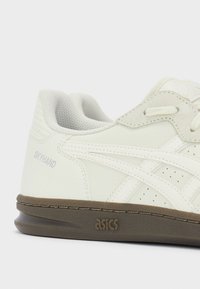 Baskets Asics Skyhand blanches avec accents en daim beige, côtés perforés et semelle marron, vues de côté sur fond blanc.