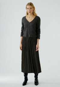 Cardigan gris texturé avec des boutons, associé à une jupe maxi plissée noire et des bottes noires longues, créant une tenue élégante en couches.