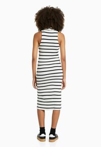 Robe blanche à dos côtelé avec des rayures verticales noires, col montant et sans manches. Des baskets noires et des chaussettes mi-mollet complètent le look.