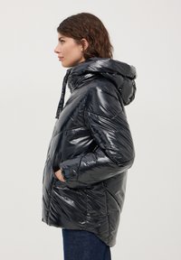 Giacca puffer nera con colletto alto, tasche laterali e texture lucida. Presenta un cappuccio con cordoncino e dettagli cuciti.