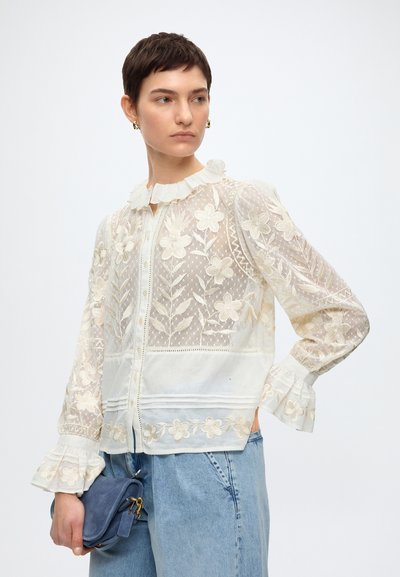 Femme aux cheveux courts portant un chemisier en dentelle florale blanche avec des poignets et un col à volants, un jean bleu, tenant un petit sac à main bleu.