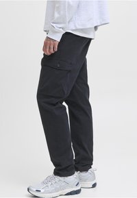 Czarne spodnie cargo z bocznymi kieszeniami, wykonane z bawełny, z prostym krojem i gładką teksturą. Noszone z szarymi butami sportowymi.