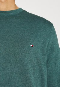 Ingrandimento di una persona che indossa un maglione di lana verde con un piccolo logo rettangolare rosso, bianco e nero sul petto.