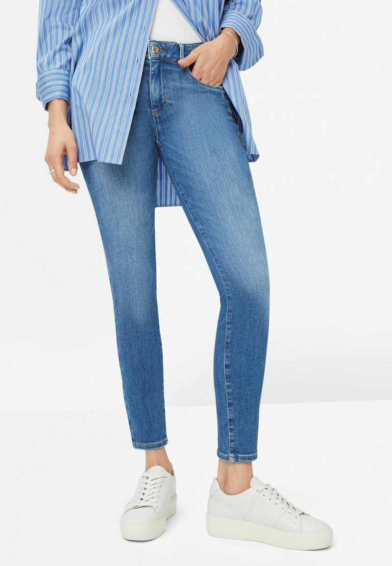 Jeans ajustados azules de mezclilla, con bolsillos frontales, cremallera y cierre de botón, combinados con zapatillas de plataforma blancas.