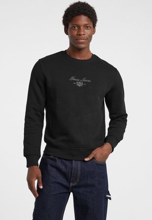 Felpa nera realizzata in misto cotone, con polsini e orlo a coste, caratterizzata dal logo ricamato "Guess Jeans" sul petto.